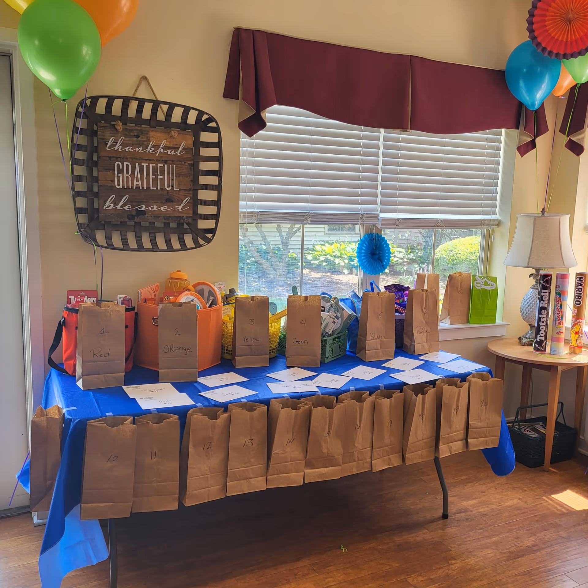 A table covered with a blue tablecloth displays multiple brown paper bags labeled with numbers and colors. Behind the table, there are colorful balloons and a wall decoration with the words 'thankful, grateful, blessed.' A window with closed blinds lets in natural light, and a small round table nearby holds large candy containers and a lamp.