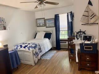 A cozy bedroom with a single bed dressed in white and blue bedding. The room features wooden flooring, a ceiling fan, two framed pictures on the wall above the bed, a window with blue curtains, a small desk with a chair, and nautical-themed decorations including a sailboat wall hanging and a ship's wheel.