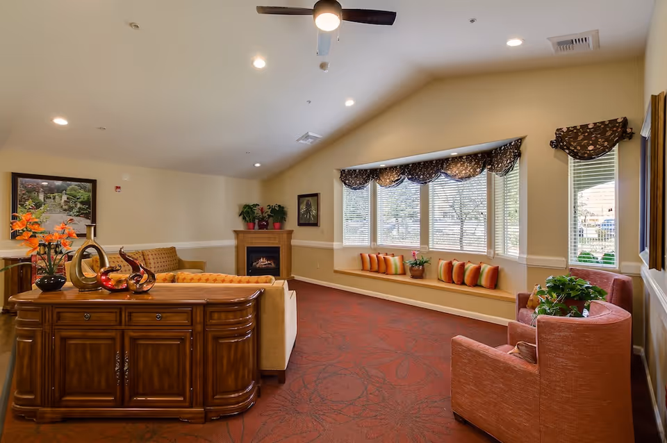 Bright, comfortable memory care common room with sofas, armchairs, a fireplace, a decorative console, and a window bench with cushions.