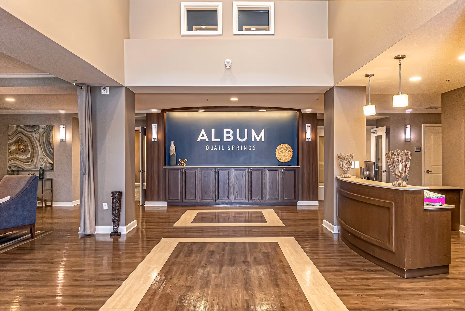 Interior view of the reception area at Album Quail Springs featuring a wooden reception desk on the right, a decorative wall with the facility name in the center, and a seating area partially visible on the left. The space has polished wooden floors with light-colored inlays and warm lighting.