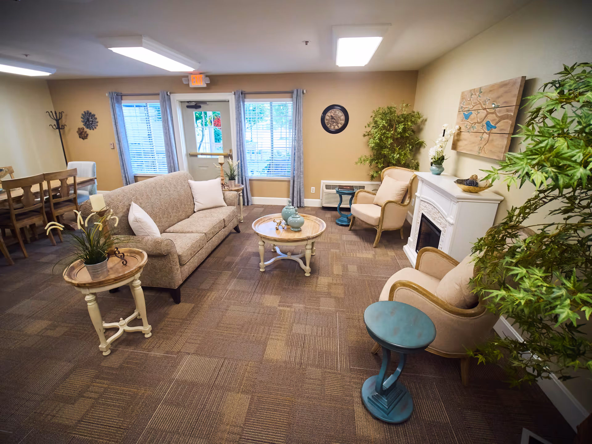A cozy living room area with a beige patterned sofa, two beige armchairs, a white fireplace, and several small tables with decorative items. The room has large windows with gray curtains, a wall clock, and green plants adding a touch of nature. In the background, there is a wooden dining table with chairs.