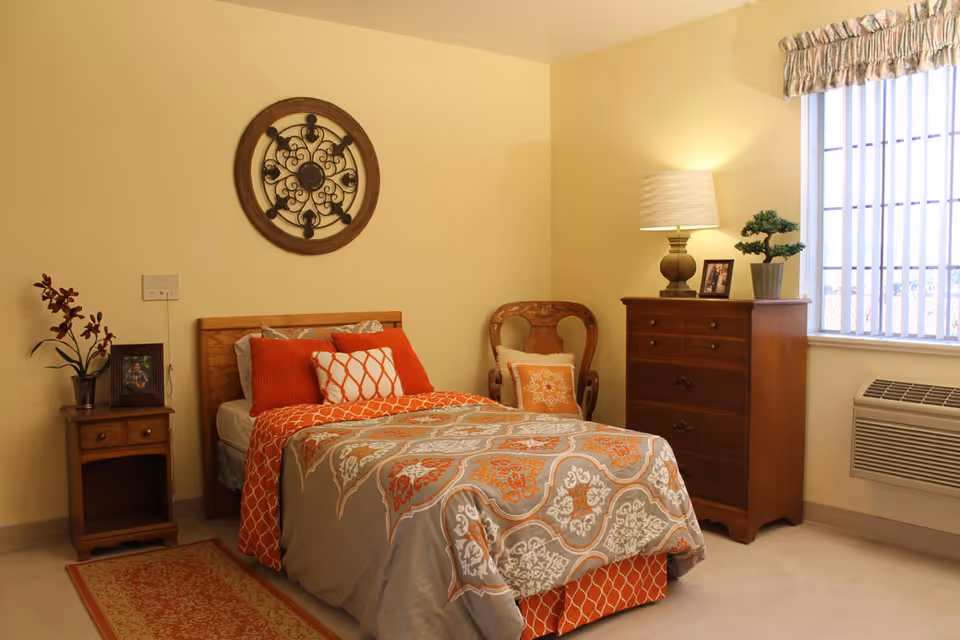 A cozy bedroom with a single bed dressed in a patterned orange and gray comforter and multiple pillows. Next to the bed is a wooden nightstand with a potted plant and a framed photo. A decorative round wall piece hangs above the bed. There is a wooden chair with a cushion beside a wooden dresser topped with a lamp, a small potted plant, and a framed photo. A window with vertical blinds and a floral valance lets in natural light, and an air conditioning unit is installed below the window.