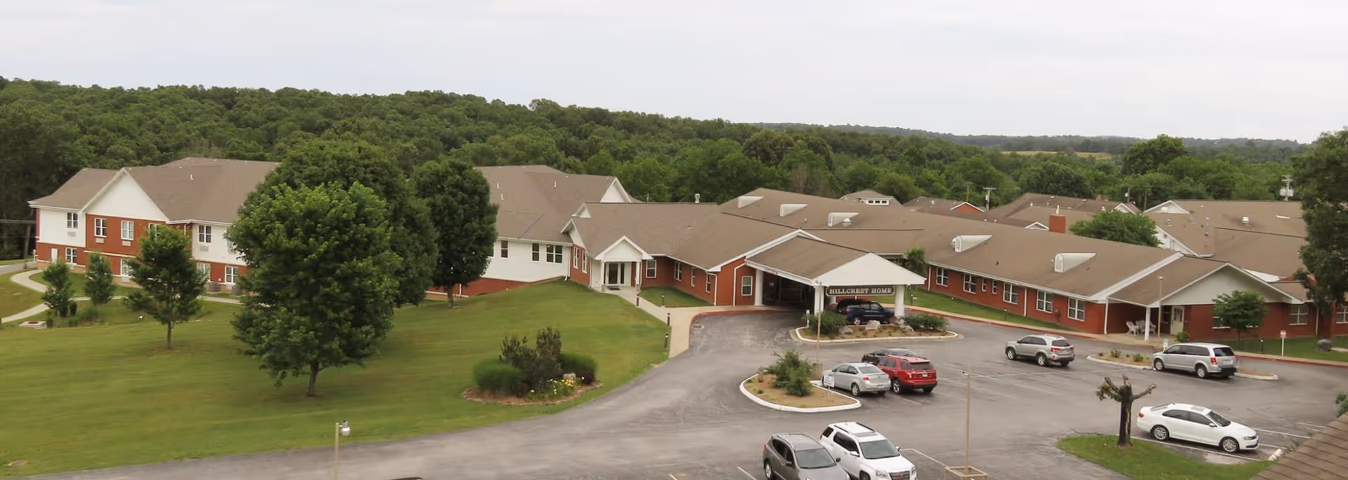 Aerial view of Hillcrest Home, a large single-story senior living facility with red brick and white siding exterior, surrounded by green lawns and trees. Several cars are parked in the driveway and parking area in front of the building. The facility is set against a backdrop of dense forest and open sky.