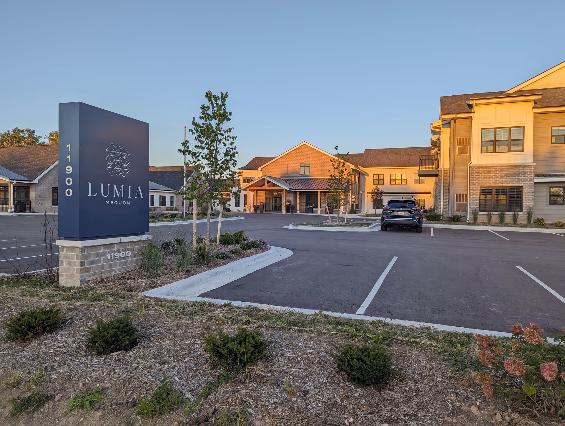 Exterior view of Lumia Mequon Assisted Living & Memory Care facility during daylight, showing a large sign with the facility name and address 11900, a parking lot with a single parked car, landscaped areas with small bushes and trees, and the multi-story building with windows and a covered entrance.