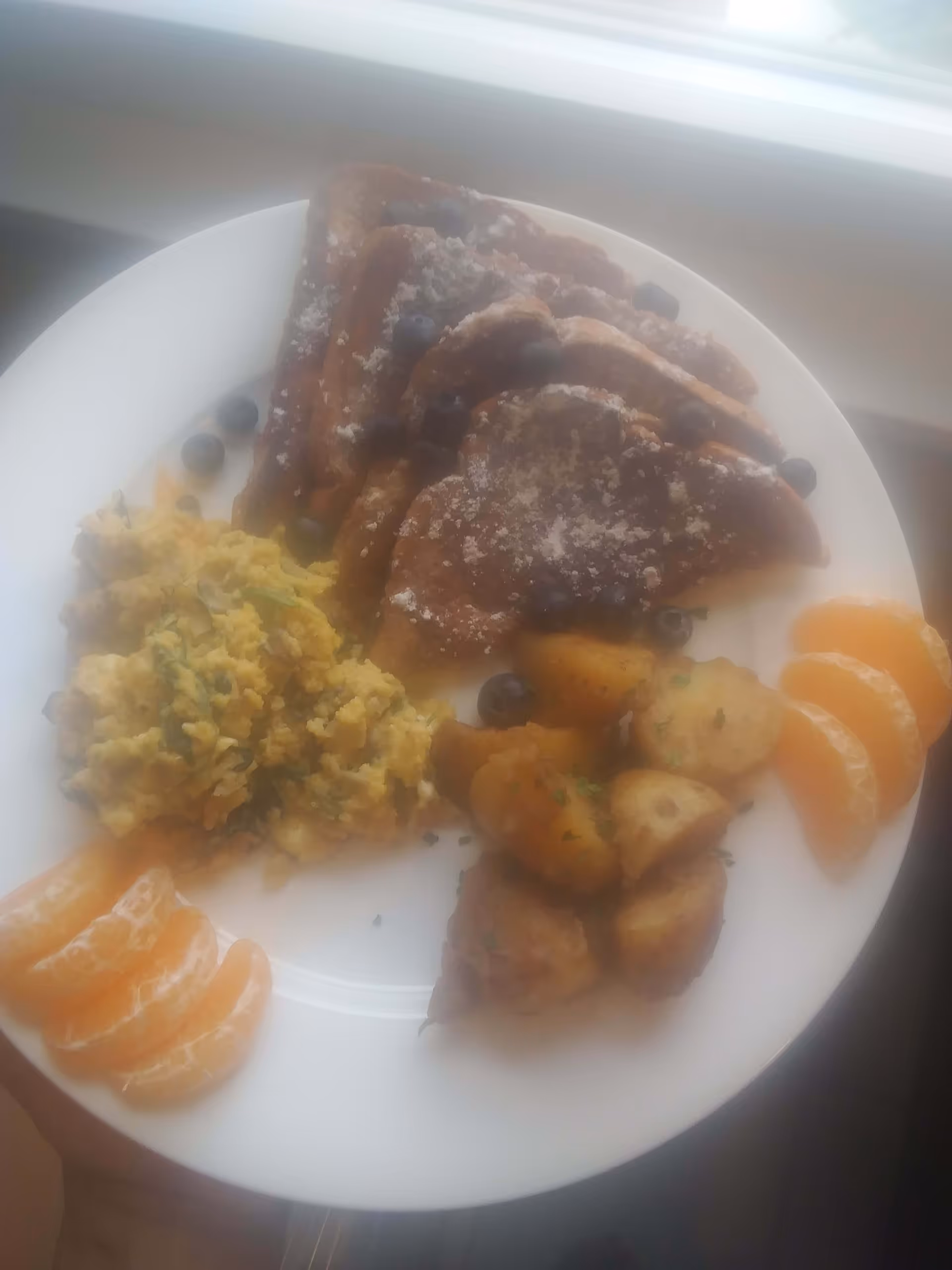 A white plate with a breakfast meal consisting of scrambled eggs, roasted potatoes, slices of orange, and French toast topped with powdered sugar and blueberries.