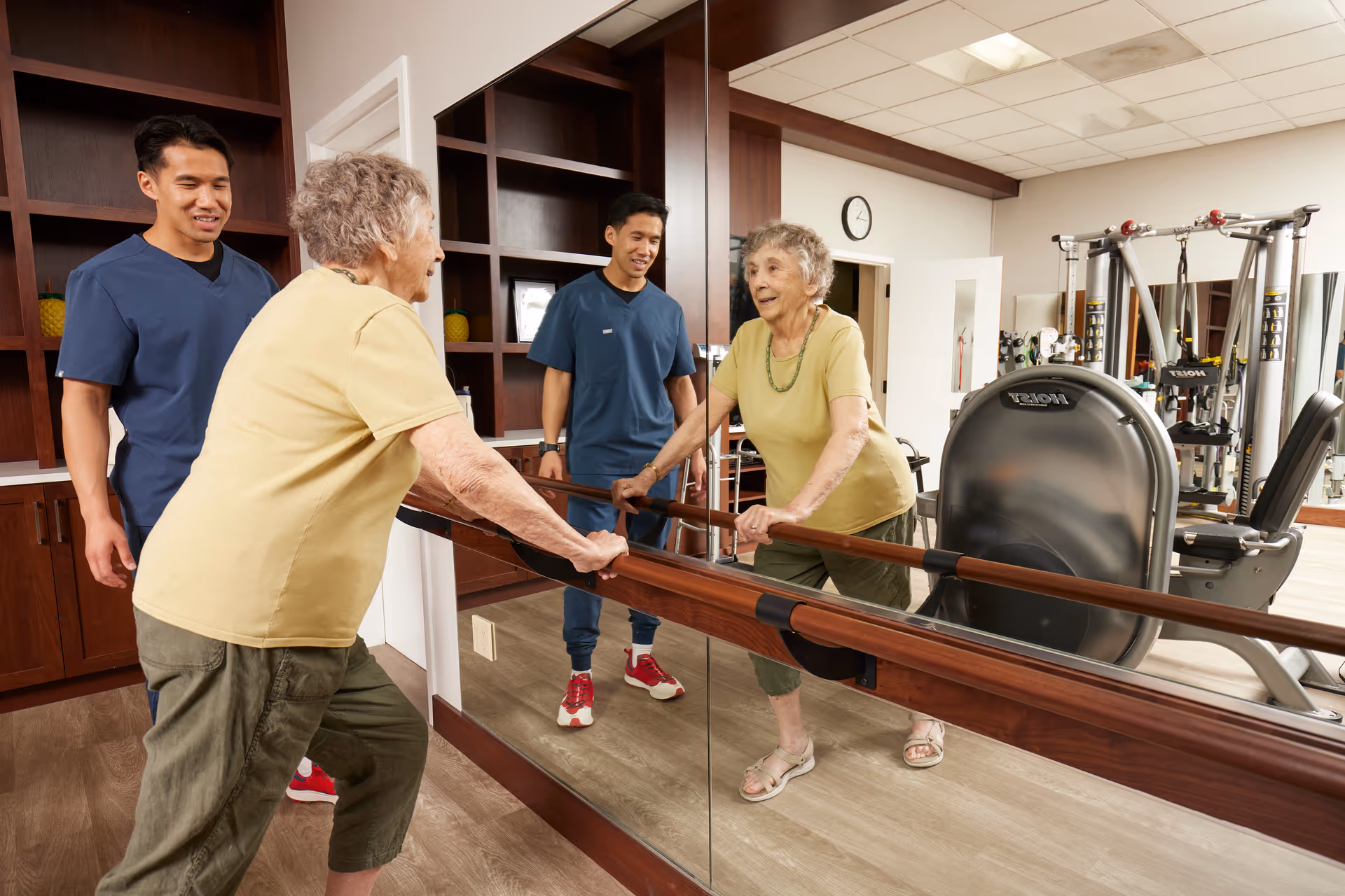 An elderly woman in a yellow shirt and green pants is holding onto a wooden ballet barre in front of a large mirror, performing a stretching exercise. A young male caregiver in blue scrubs stands nearby, smiling and observing her. The room has wooden shelves, exercise equipment, and a clock on the wall.