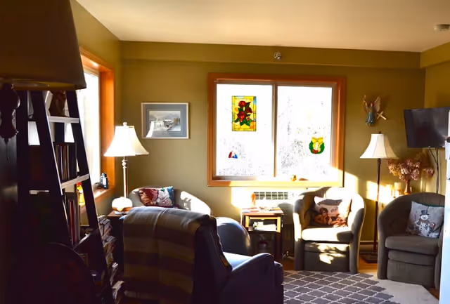 A cozy living room with three armchairs arranged around a small table under a window with stained glass decorations. The room has warm lighting from two floor lamps, a patterned rug on the floor, a wall-mounted TV, and various decorative items including framed artwork and a small angel wall hanging.