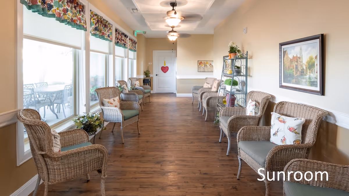 Bright sunroom corridor with wicker chairs arranged along both walls, large windows on the left, and hardwood floors.
