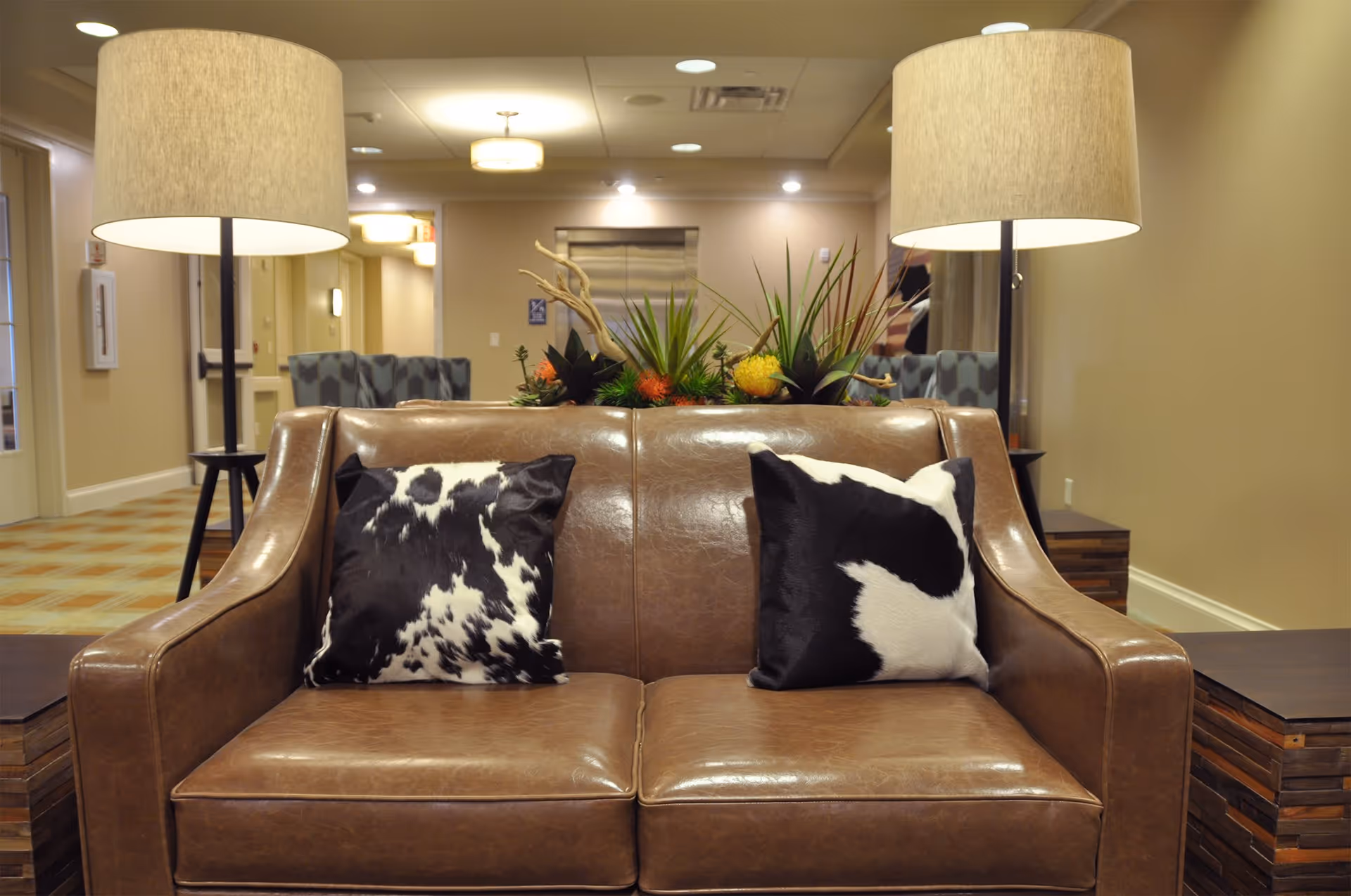 A brown leather loveseat with two black and white patterned pillows is positioned between two wooden side tables, each with a tall lamp with a beige shade. Behind the loveseat is a decorative arrangement of green plants and flowers. The background shows a hallway with beige walls, ceiling lights, and an elevator door.