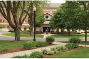 View of the entrance to a brick building surrounded by green trees, shrubs, and a well-maintained lawn with a sidewalk and a lamppost in the foreground.