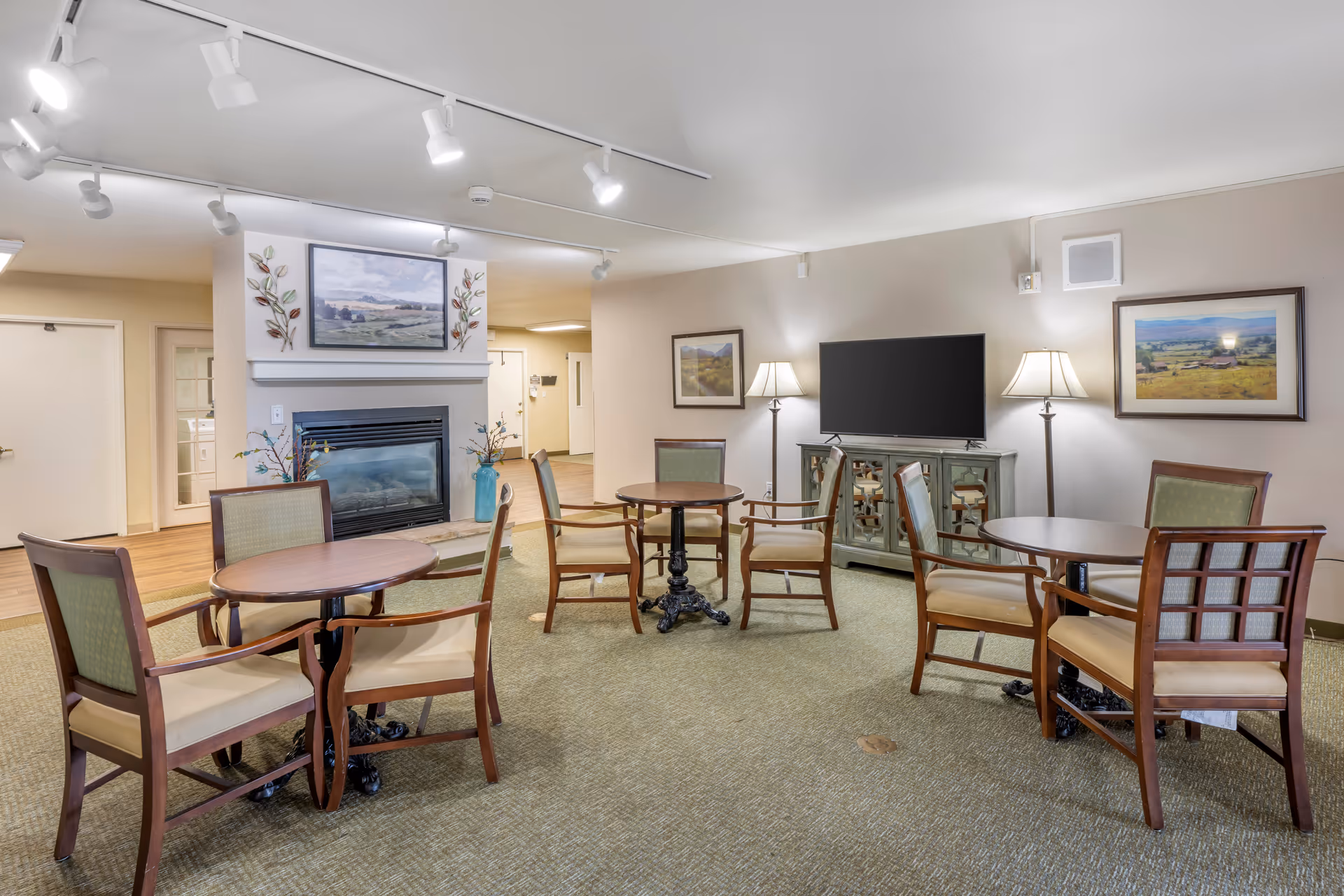 A cozy common area with three round wooden tables, each surrounded by four cushioned wooden chairs. The room features a fireplace with decorative vases and a landscape painting above it. On the right side, there is a flat-screen TV on a cabinet, flanked by two floor lamps and framed landscape artwork on the walls. The space has carpeted flooring and soft lighting from ceiling track lights.
