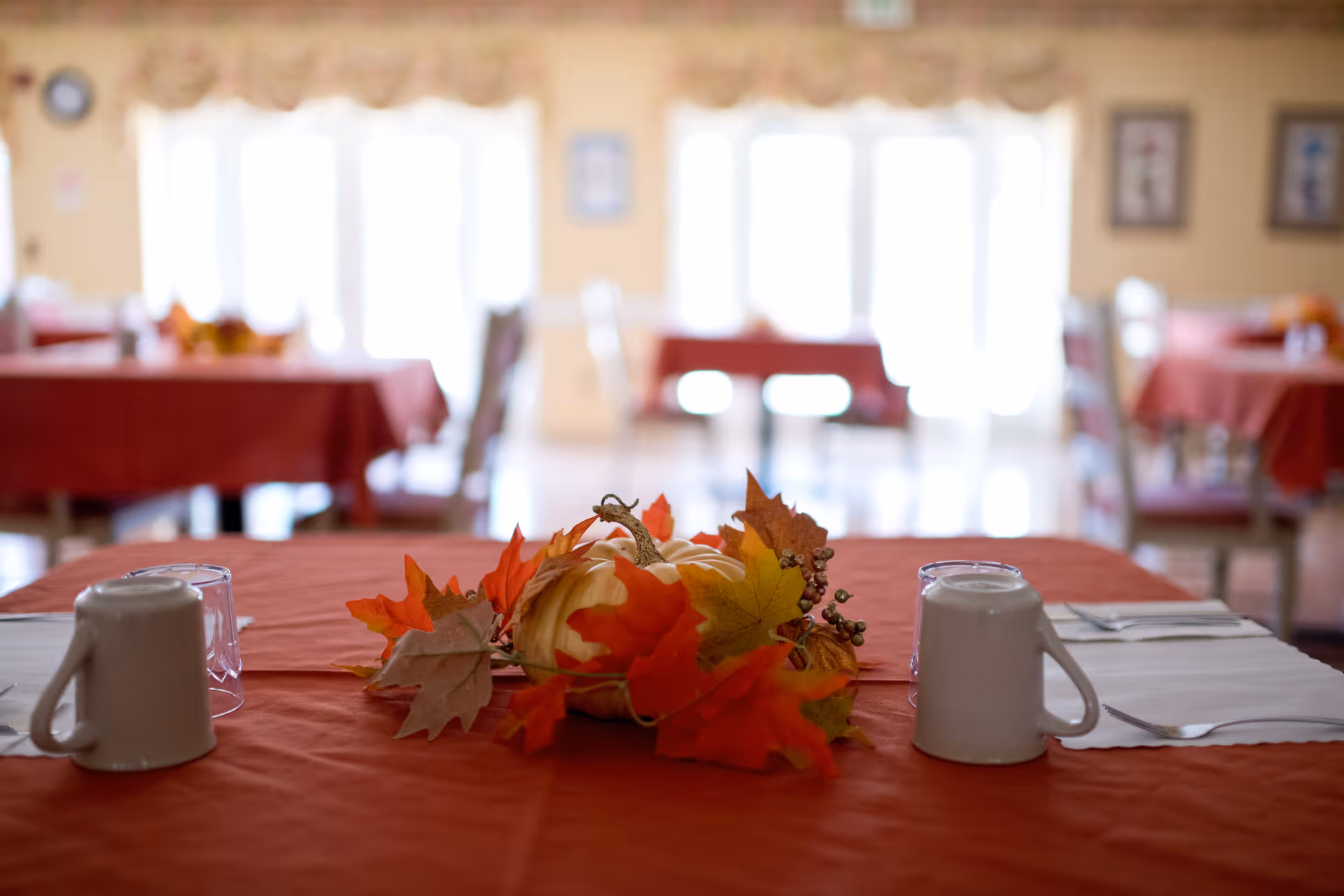 A dining table set with a red tablecloth, two upside-down white mugs, a glass, and a fall-themed centerpiece featuring a small pumpkin and autumn leaves. In the background, there are more tables with red tablecloths and chairs in a softly lit dining room with large windows.