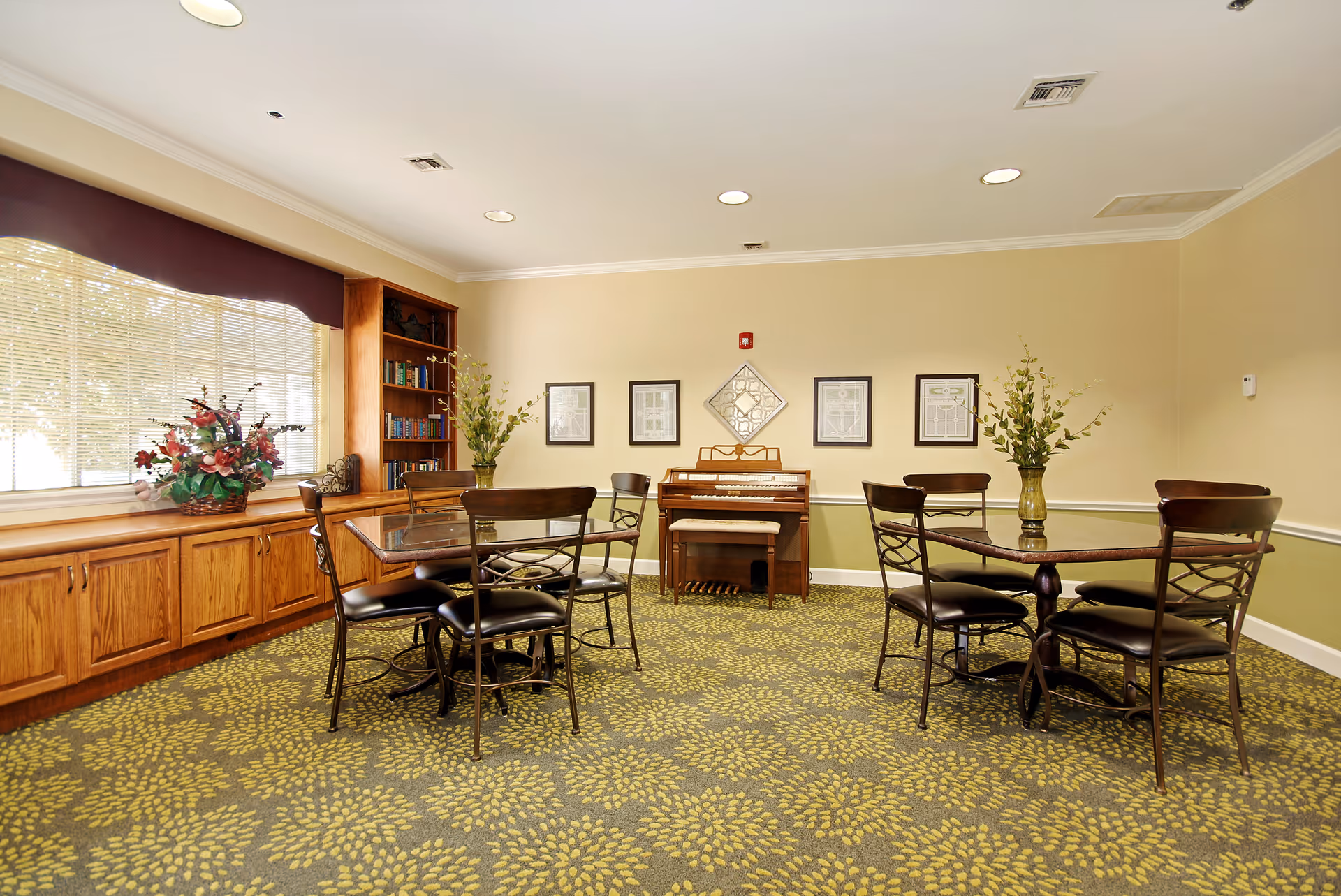 A well-lit common room with two glass-top tables, each surrounded by four metal chairs with cushioned seats. The room features a patterned carpet with green and yellow leaf designs, beige walls with a green lower half, and a large window with blinds and a dark valance. There is a wooden built-in bookshelf filled with books and decorative items, a floral arrangement on the windowsill, and a small piano against the far wall with framed artwork above it. Two vases with green plants are placed on the tables.