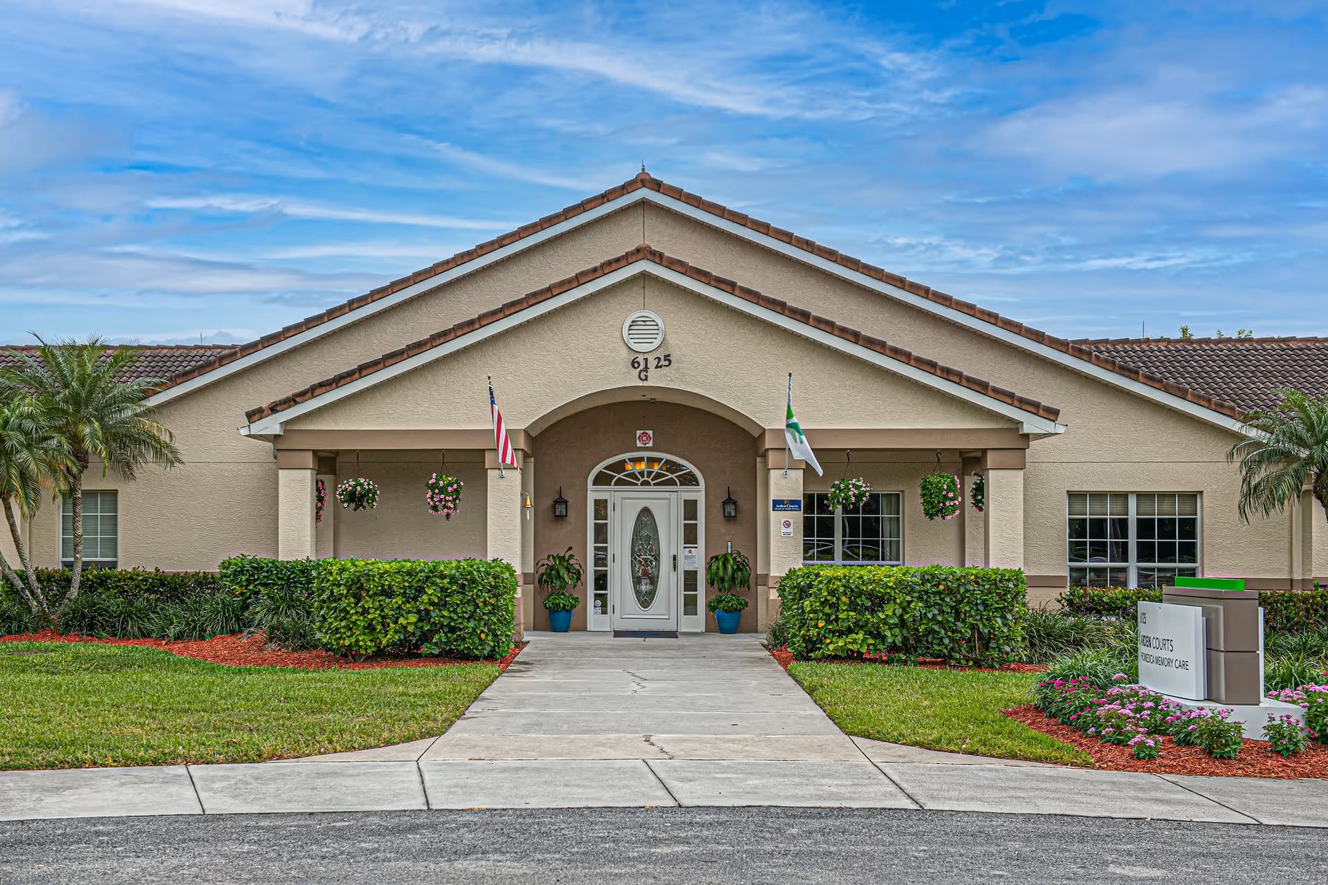 Front exterior view of a single-story senior living facility building with a beige facade and a tiled roof. The entrance features a white door with glass panels, flanked by two potted plants and hanging flower baskets. Two flags are displayed on either side of the entrance. The surrounding landscaping includes green bushes, palm trees, and a well-maintained lawn with flower beds.