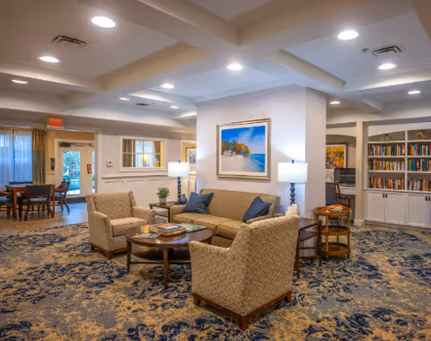 A cozy senior living facility common area with a beige sofa and two patterned armchairs arranged around a round wooden coffee table. The room features a blue and beige patterned carpet, a painting of a beach scene on the wall, two table lamps, and built-in bookshelves filled with books. In the background, there is a dining area with a table and chairs near a door leading outside.