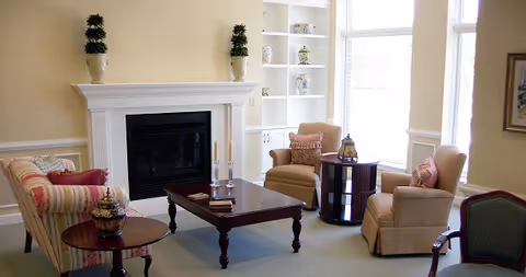 A cozy living room with a fireplace, two beige armchairs with patterned pillows, a striped sofa, a wooden coffee table with candles and books, and built-in white shelves with decorative items. Large windows allow natural light to fill the room.