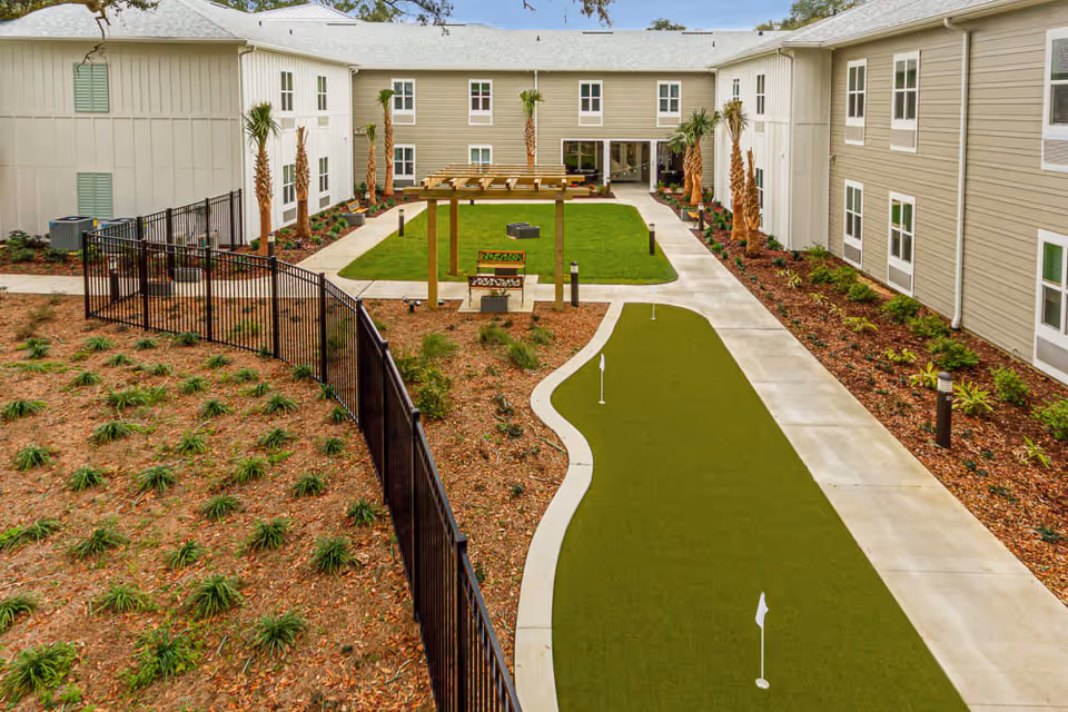 Courtyard of a senior living facility with a putting green, pergola, walkways and surrounding two-story buildings.