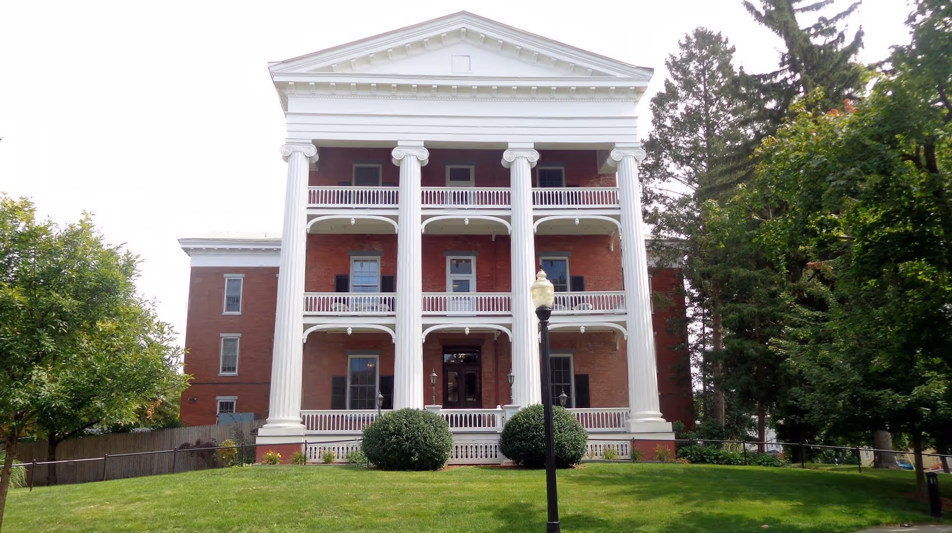 Front exterior view of a large, historic brick building with white columns and balconies, surrounded by green trees and a well-maintained lawn.