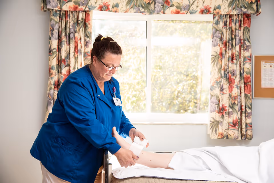 A healthcare worker in a blue uniform is wrapping a bandage around the ankle of a person lying on a bed in a room with floral curtains and a window letting in natural light.