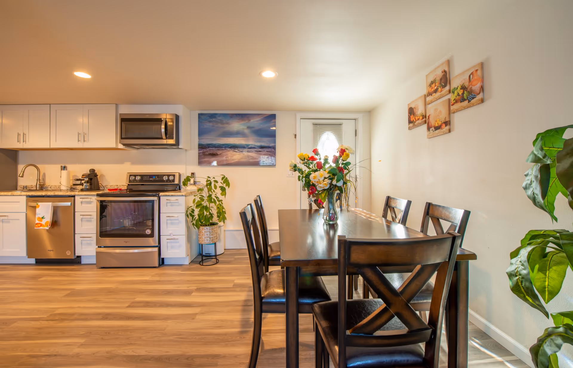 A bright and clean dining area with a dark wooden table and six matching chairs. A vase with colorful flowers is placed on the table. The room features light wood flooring and white walls decorated with framed pictures of fruit and pottery. To the left, there is a modern kitchen with white cabinets, stainless steel appliances including a microwave, stove, and dishwasher. A potted plant is placed near the kitchen area, and a door with a frosted glass window is visible in the background.