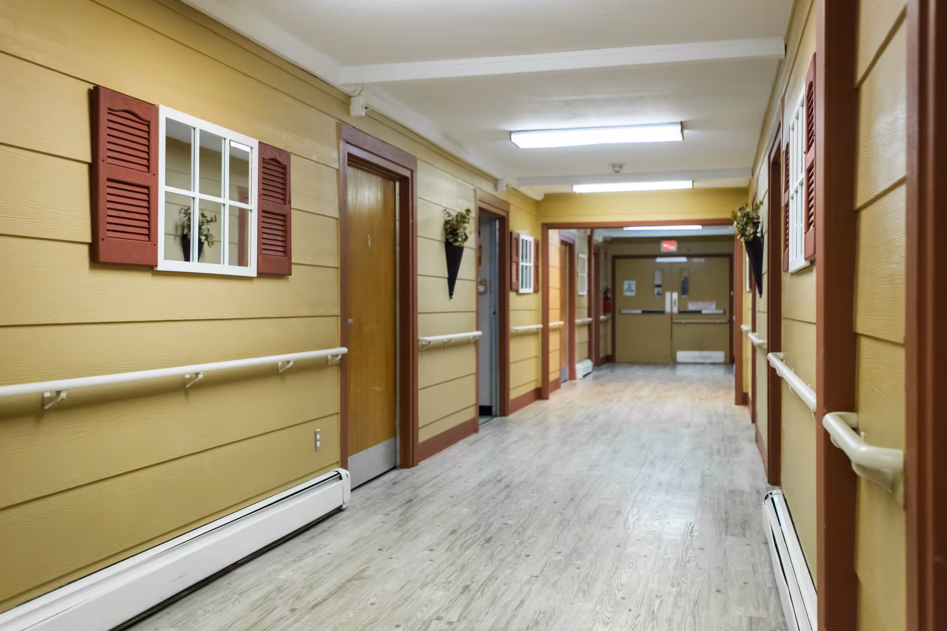 A long indoor hallway with yellow walls and wooden doors on both sides, decorated with small window-like wall hangings and flower arrangements. The floor is light-colored wood, and there are handrails along both walls. Fluorescent ceiling lights illuminate the corridor.