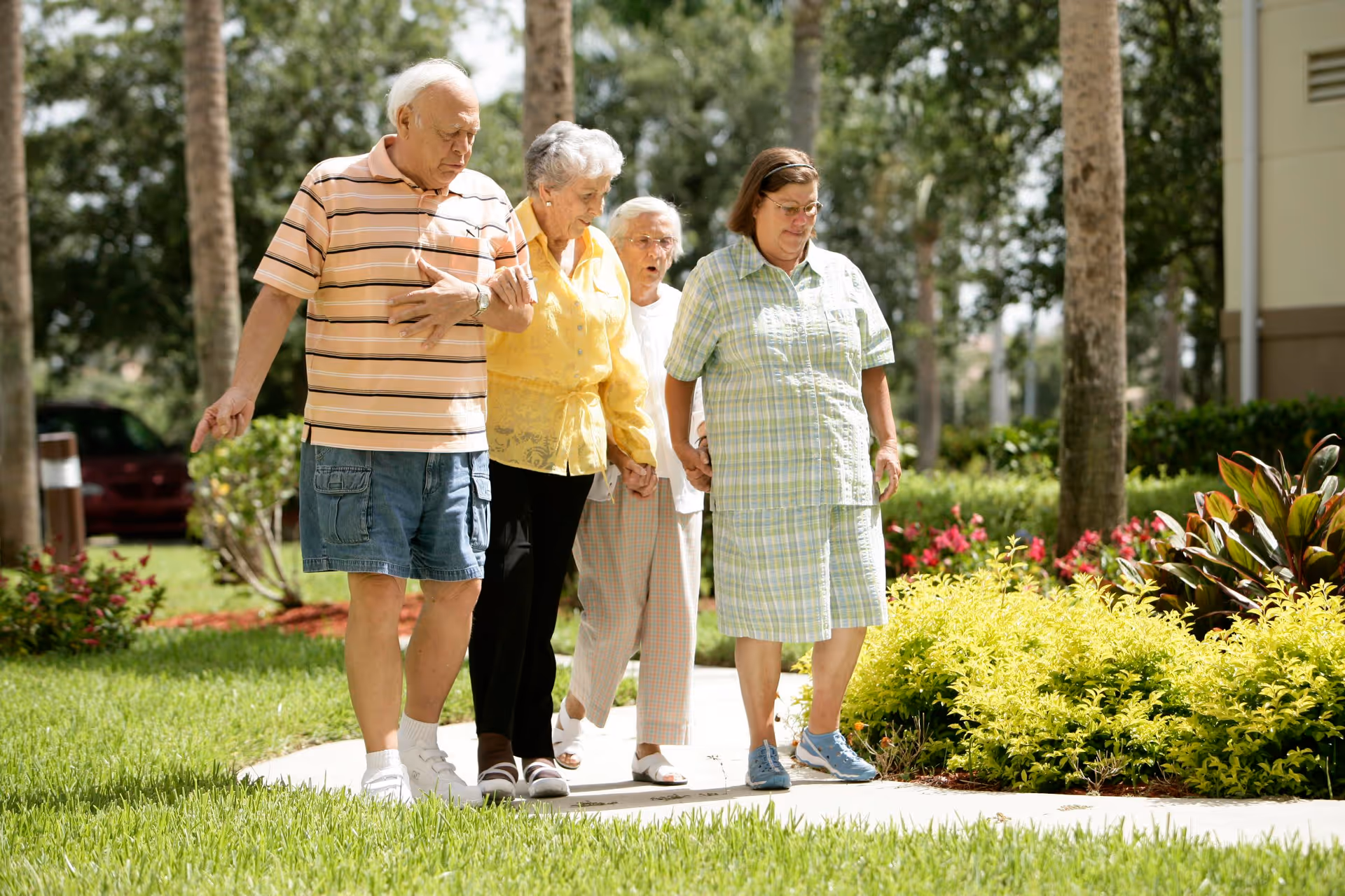 Four elderly individuals walking together on a paved path outdoors, surrounded by green grass, plants, and trees. They appear to be enjoying a sunny day in a garden or landscaped area.