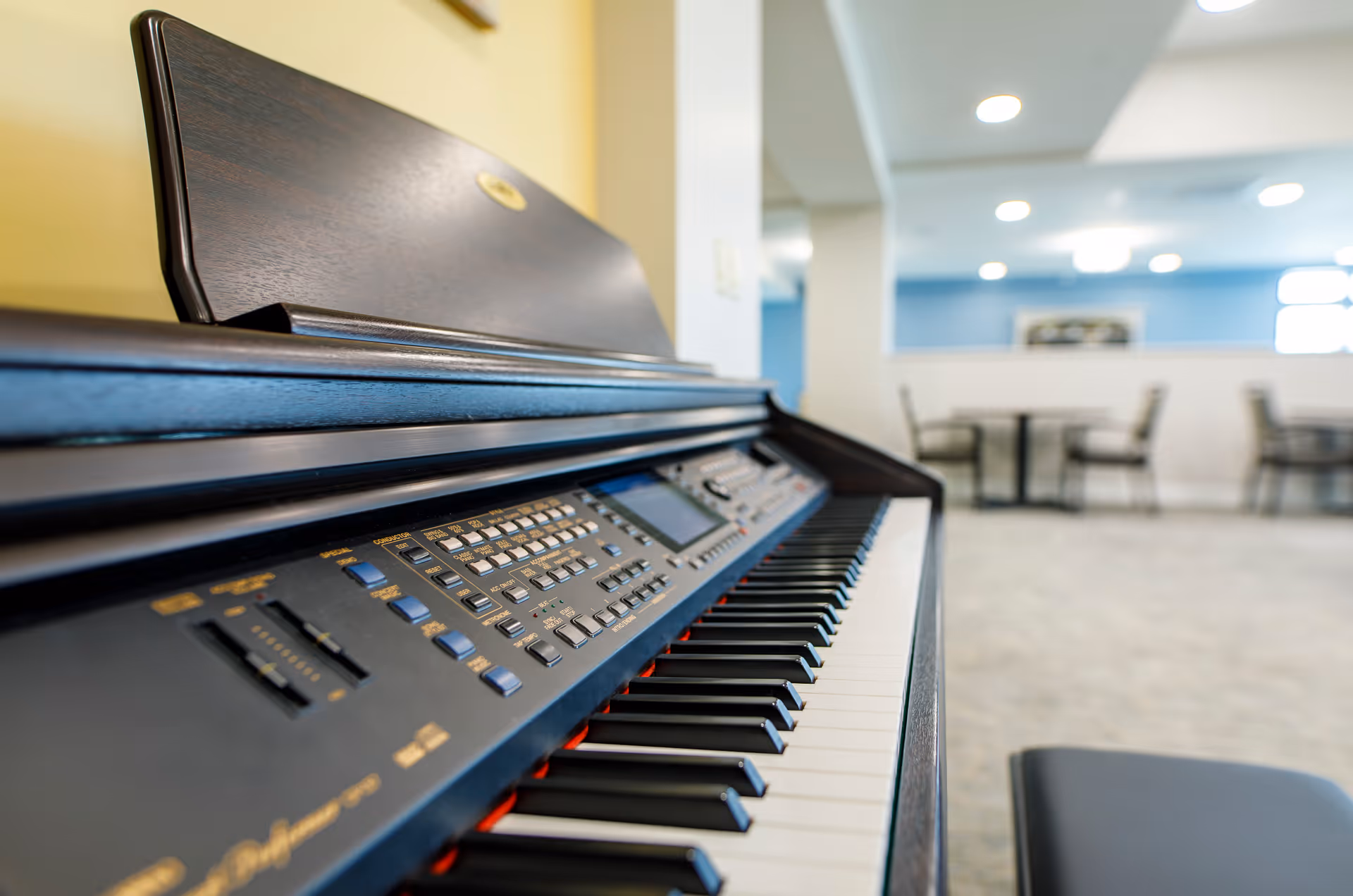 Close-up of an electronic piano keyboard in a bright communal room with tables and chairs in the background.