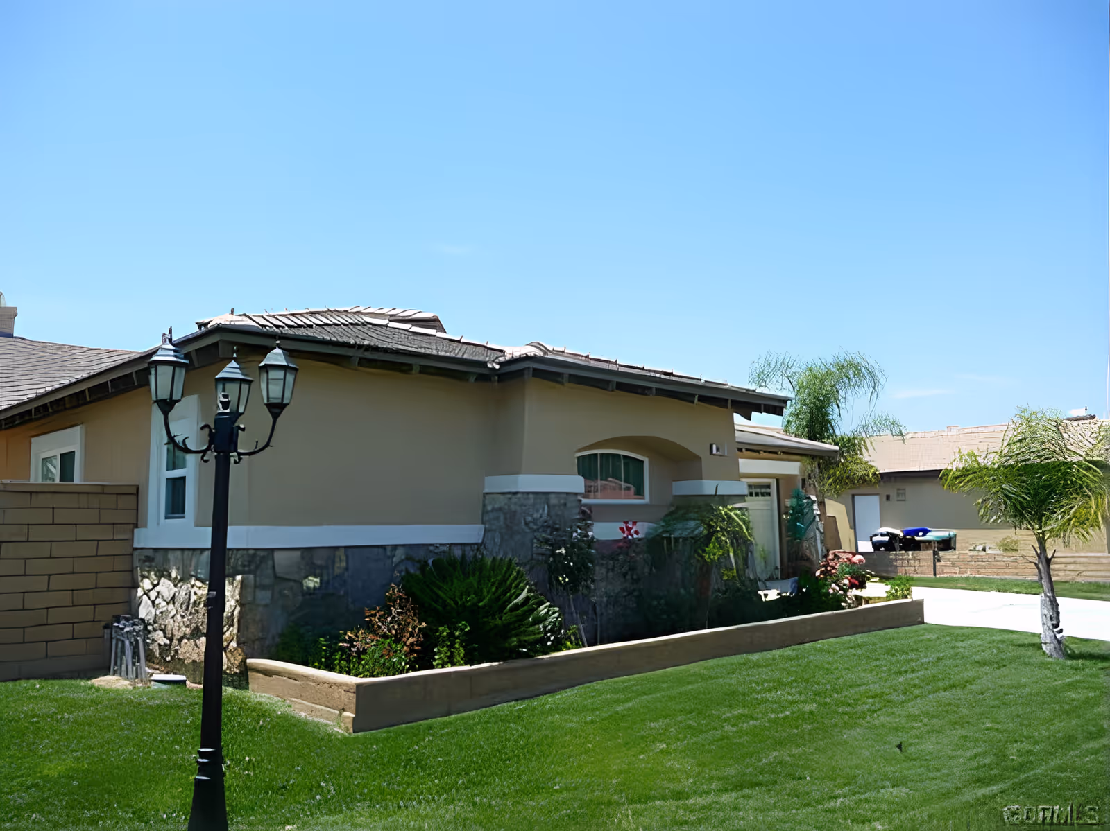 Exterior view of a single-story residential building with beige walls and stone accents, surrounded by a well-maintained green lawn and small palm trees under a clear blue sky. A black street lamp is visible in the foreground.