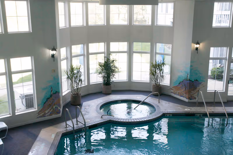 Indoor swimming pool area with a circular hot tub in the center, surrounded by large windows letting in natural light. There are potted plants placed near the windows and wall-mounted lantern-style lights. The walls feature murals of a lighthouse and coastal scenery.