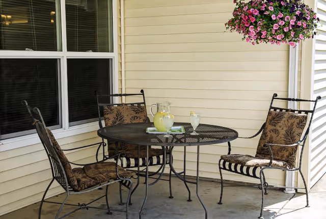 Outdoor patio area with a round metal table and three cushioned metal chairs. On the table, there is a pitcher of lemonade, a glass of lemonade with a lemon slice, and a book. A hanging basket with pink and purple flowers is visible above the table. The patio is adjacent to a building with cream-colored siding and a window with closed blinds.