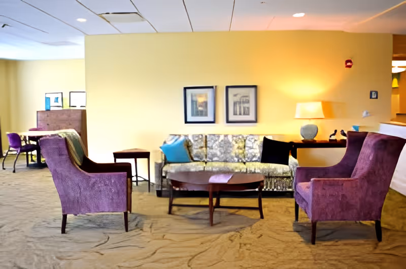 A cozy living room area with a yellow wall, a patterned sofa with blue and black cushions, two purple armchairs, a wooden coffee table, and a side table with a lamp. In the background, there is a small table with chairs and framed pictures on the wall.