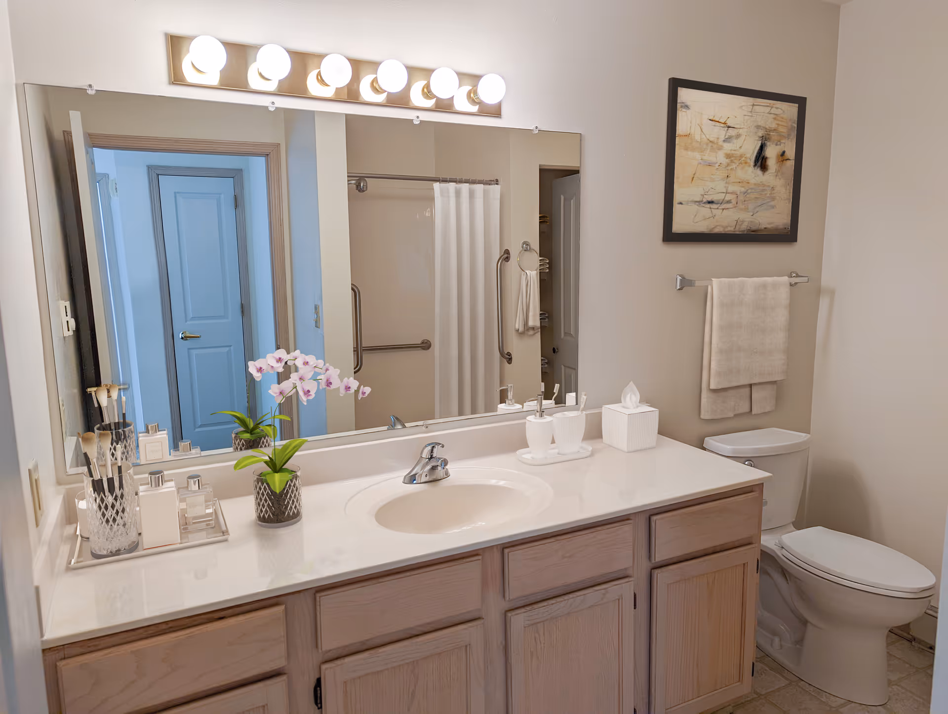 Bright bathroom with a vanity countertop and sink, large mirror and lights, toilet, and a shower visible in the mirror.