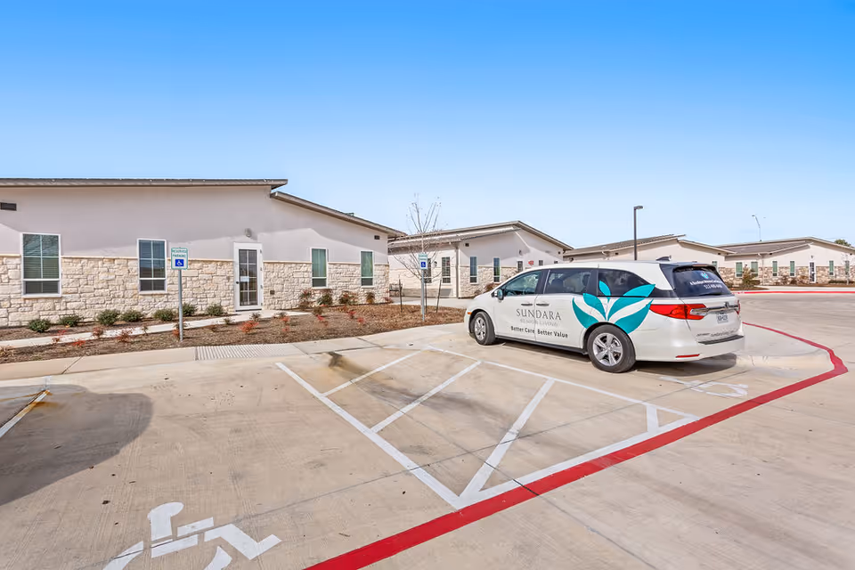 Parking lot and building front of a single-story Sundara Senior Living facility with a branded Sundara van parked in a handicapped space under a clear sky.