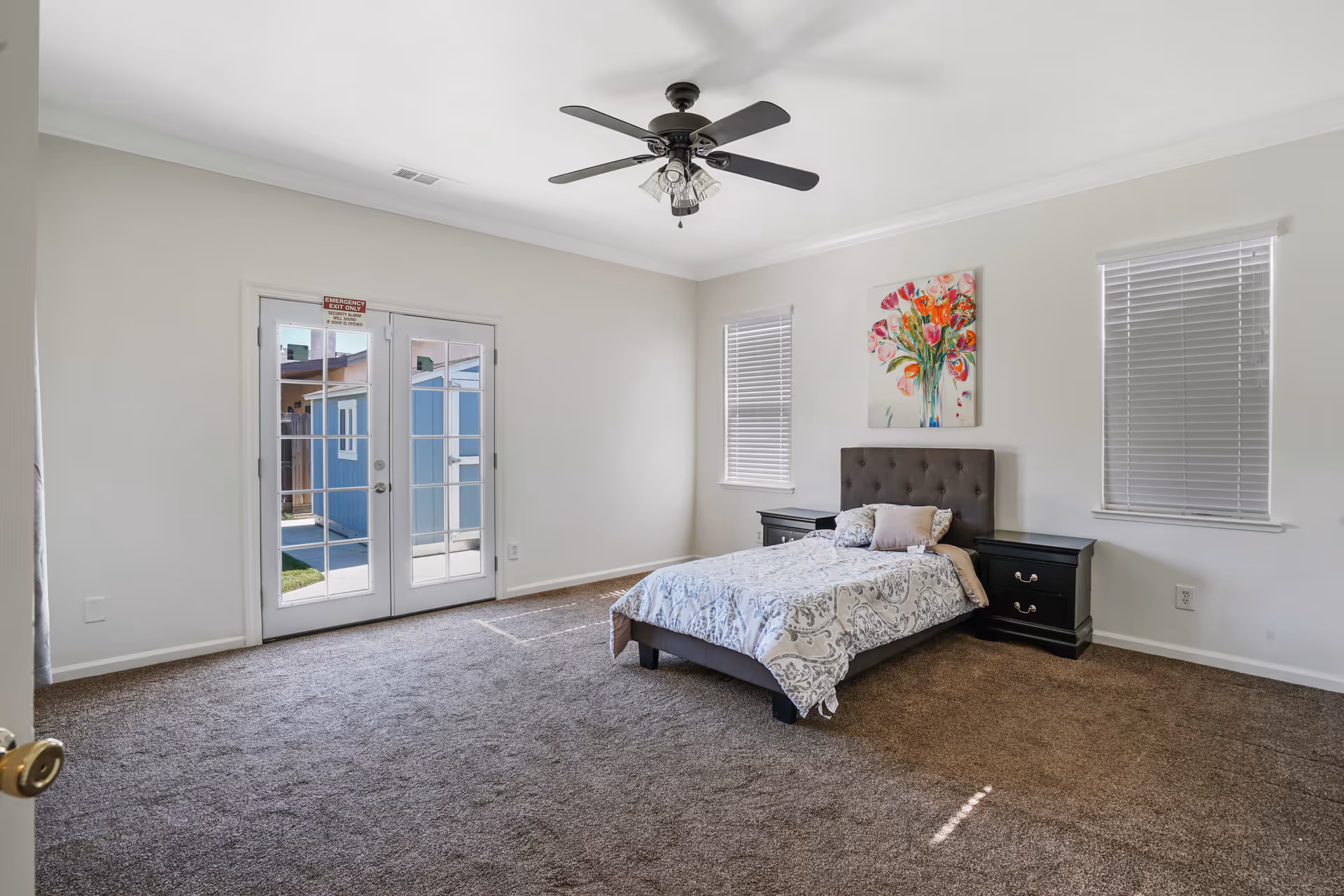 A spacious bedroom with a bed centered against the wall, flanked by two black nightstands. The bed has a patterned comforter and several pillows. Above the bed hangs a colorful painting of flowers. The room has beige carpet, white walls, two windows with blinds, and a ceiling fan with lights. French doors lead outside to a patio area with a blue shed visible.
