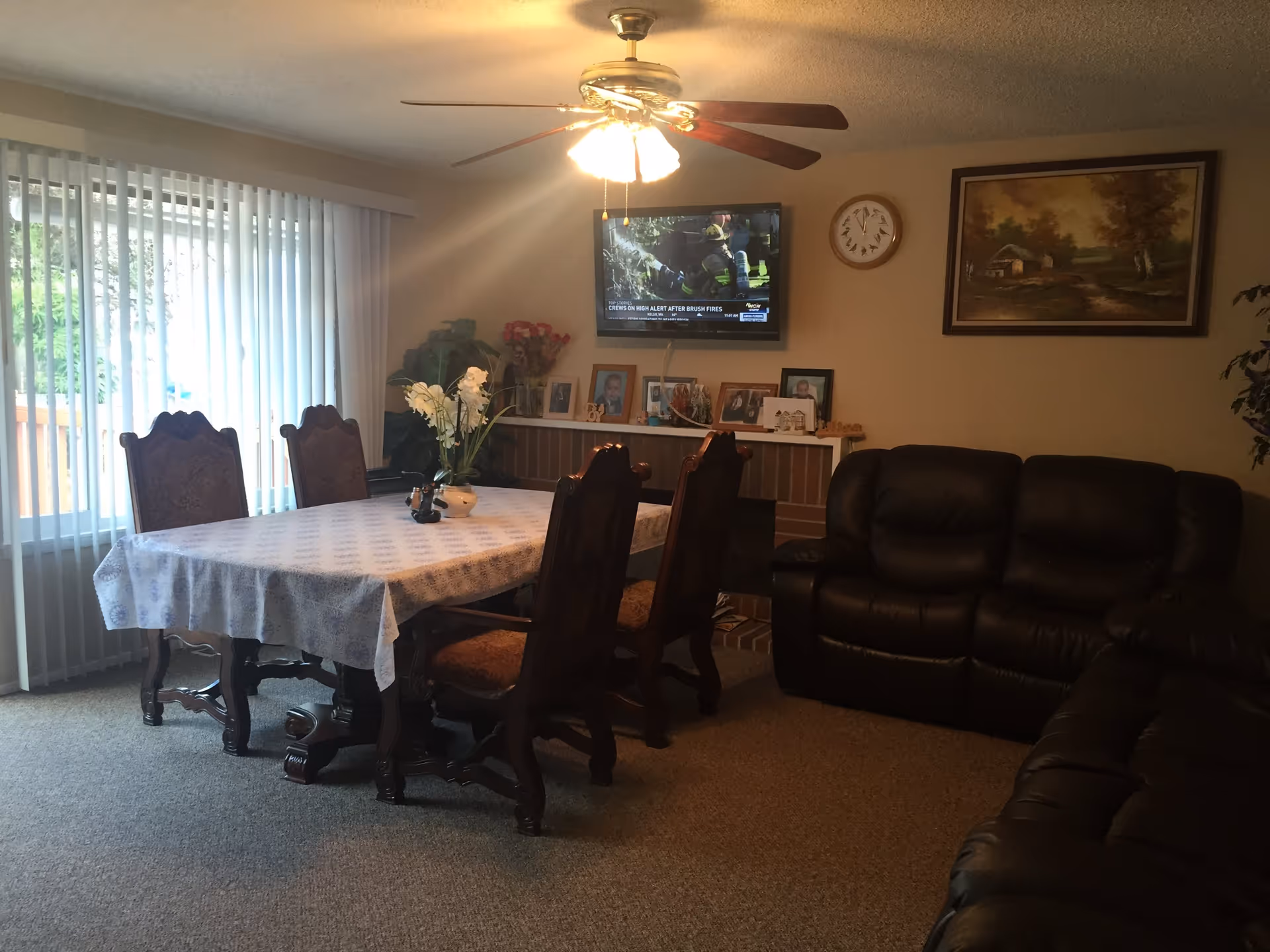 A cozy living and dining area with a table covered by a white patterned tablecloth surrounded by four wooden chairs. A ceiling fan with lights is above the table. To the right, there are two black leather sofas. On the wall, a flat-screen TV is mounted above a shelf displaying framed photos and decorative items. A clock and a framed painting hang on the wall. Vertical blinds cover a sliding glass door leading outside.