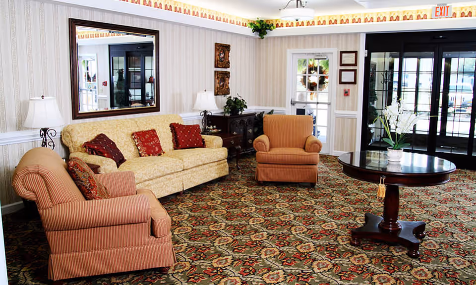 A cozy living room area with a patterned carpet, a yellow sofa with red and brown cushions, two striped armchairs, a round wooden table with a white flower vase, two table lamps, a large wall mirror, and a glass door leading outside.