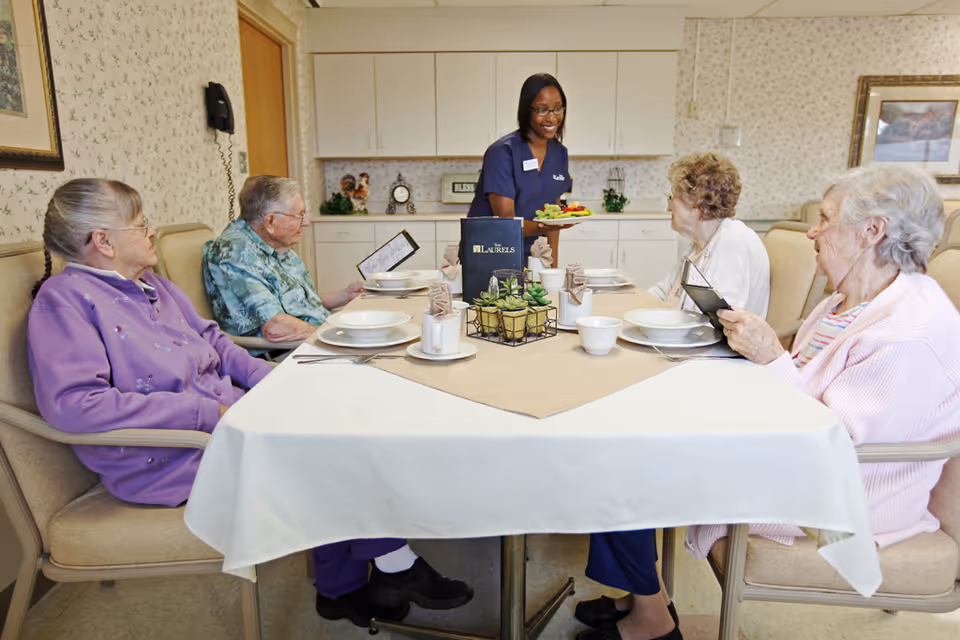 A staff member serves food to four elderly residents seated around a dining table in a communal dining room.