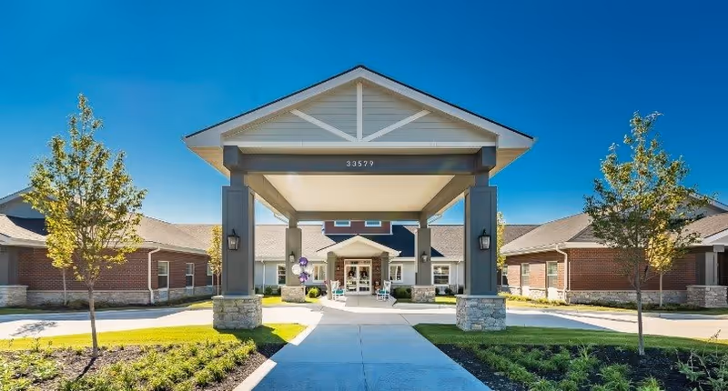 Front exterior view of Provision Living at Livonia facility with a covered entrance supported by stone and gray pillars, a walkway leading to the main doors, landscaped greenery, and clear blue sky.