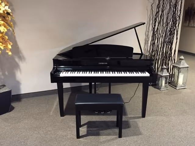 A black Yamaha grand piano with its lid open, accompanied by a matching black piano bench, placed on a carpeted floor against a plain light-colored wall. To the right of the piano are two decorative lanterns and tall, thin branches arranged vertically. To the left, there is a potted plant with yellow leaves.