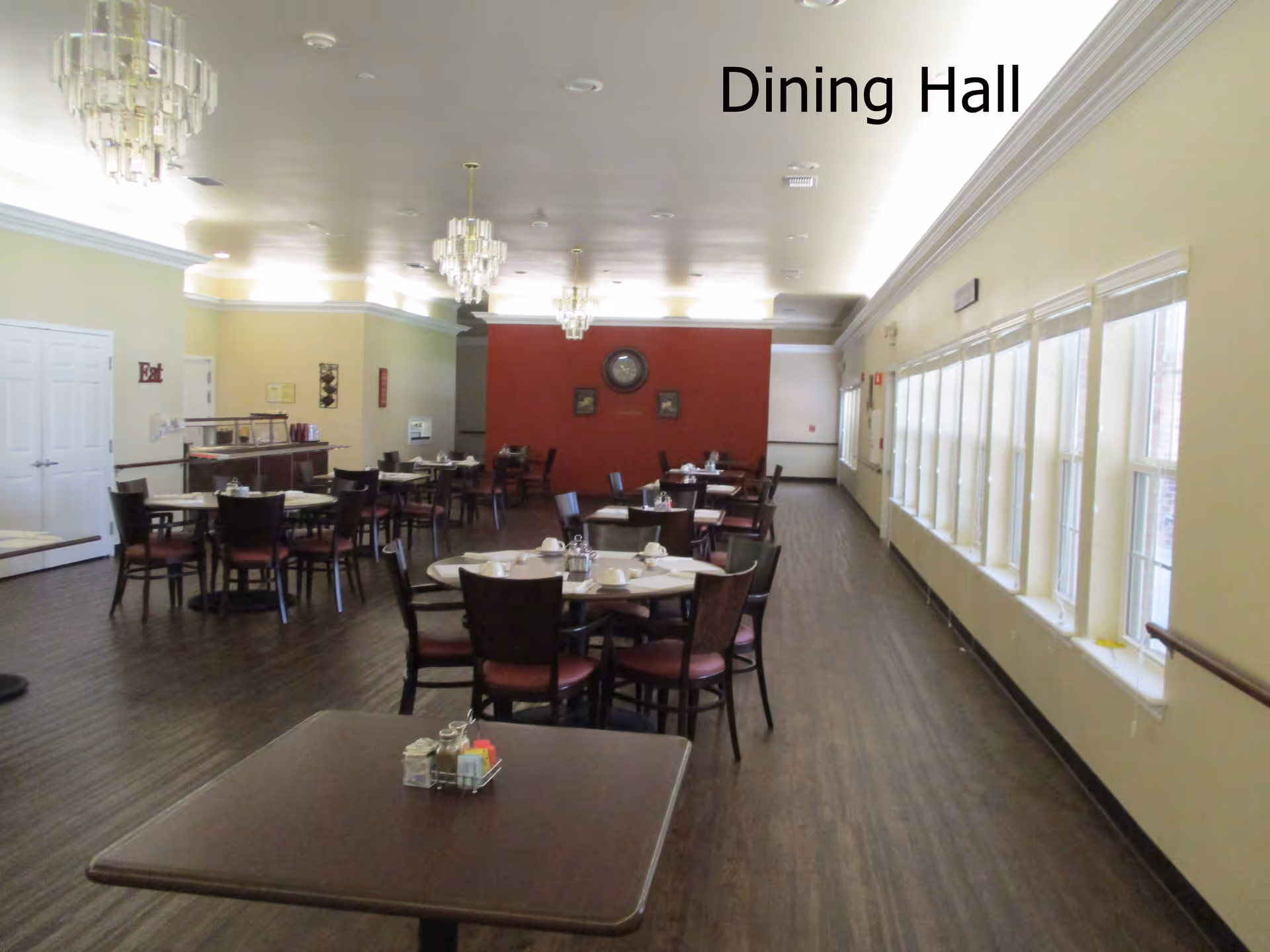 Spacious dining hall with multiple round and square tables, chairs, chandeliers, and windows along the right wall.
