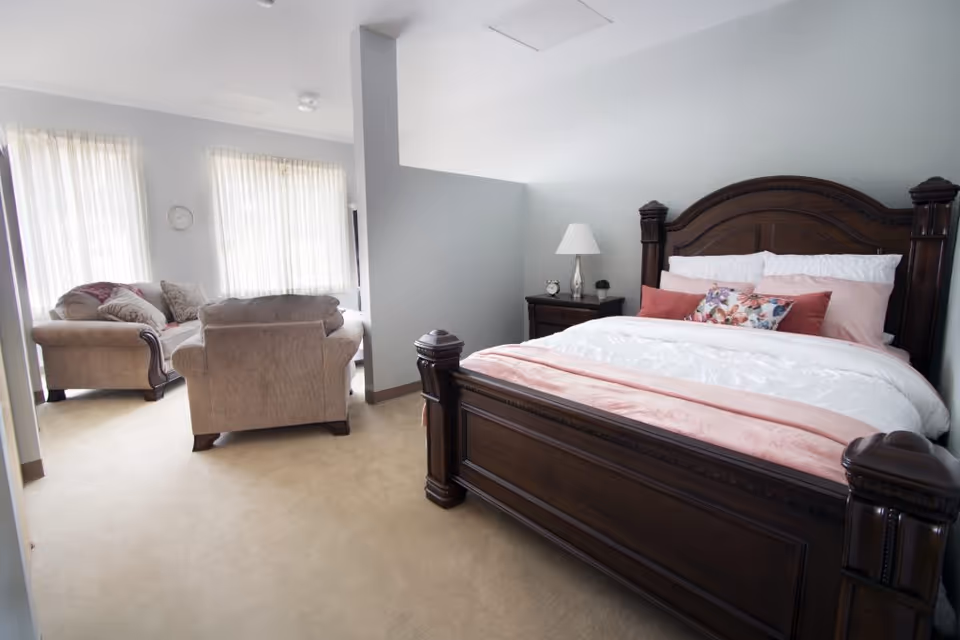 A bright bedroom with a large dark wooden bed featuring white and pink bedding and decorative pillows. Next to the bed is a nightstand with a lamp and small clock. The room has beige carpet and a partial wall separating the sleeping area from a sitting area with two beige armchairs and a loveseat near large windows with sheer white curtains.
