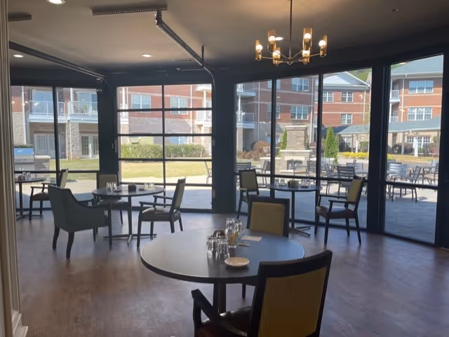 A dining area with round tables and chairs arranged inside a room with large glass windows and doors that open to an outdoor patio. The room has wooden flooring and a modern chandelier hanging from the ceiling. Outside, there are more tables and chairs, a fireplace, and a multi-story brick building in the background.
