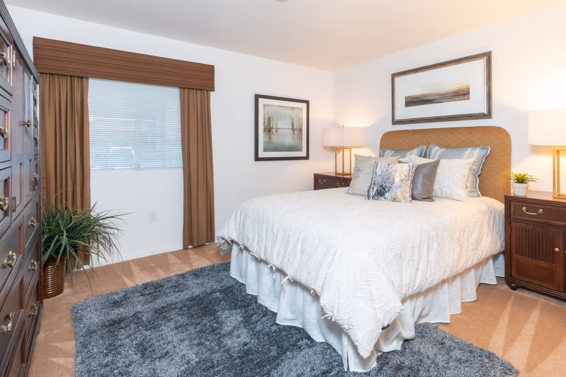 A neatly arranged bedroom with a large bed covered in white bedding and multiple decorative pillows. The room features two wooden nightstands with lamps on each side of the bed, a framed artwork above the headboard, a large wooden dresser on the left, a window with brown curtains, a green potted plant, and a gray area rug on the carpeted floor.