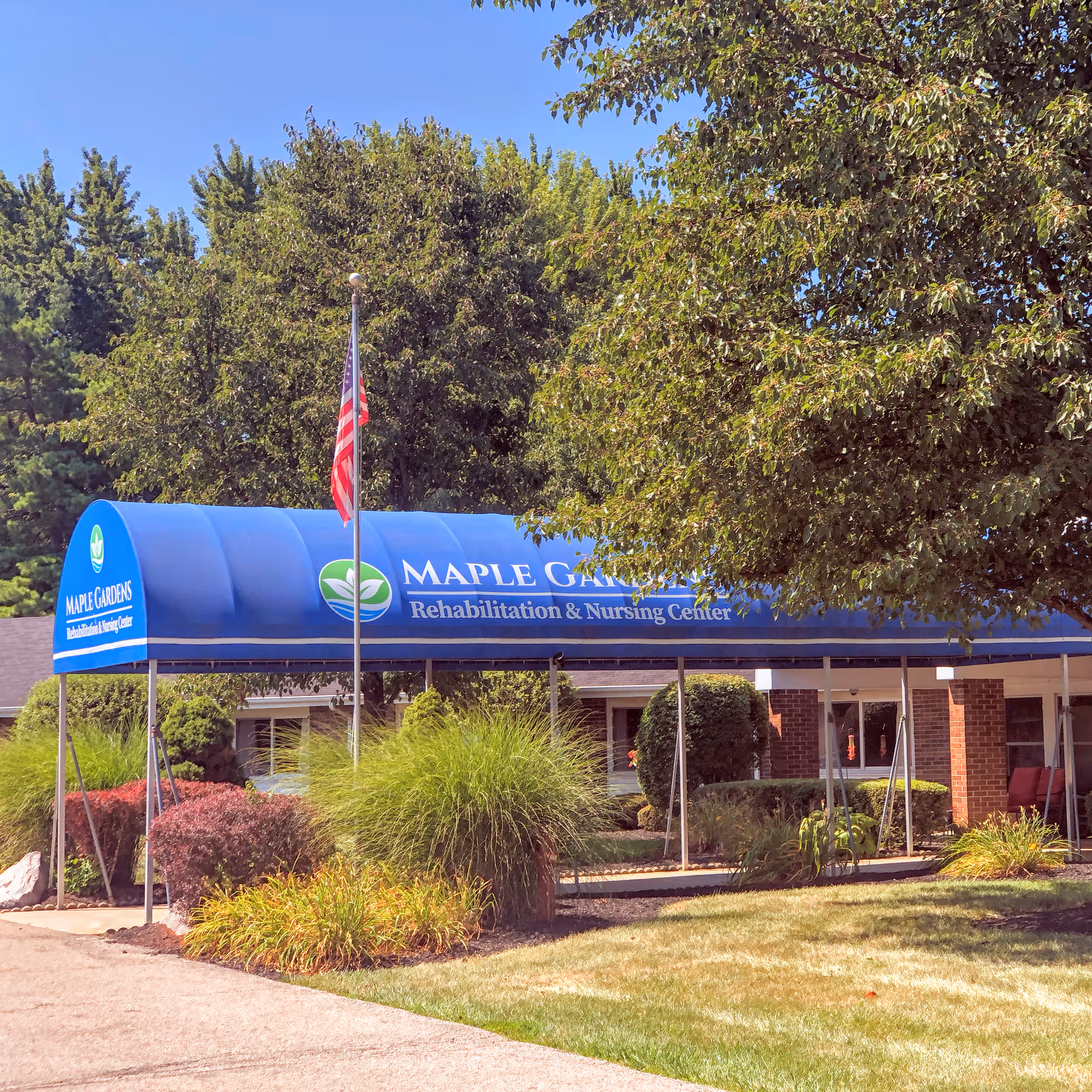Entrance of Maple Gardens Rehabilitation and Nursing Center with a blue canopy displaying the facility name, surrounded by green trees and bushes, and an American flag on a flagpole.