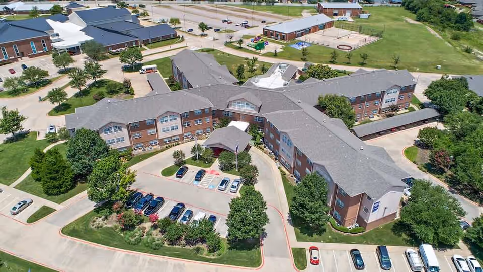 Aerial view of Morada Cedar Hill senior living facility showing a large, multi-wing building with a gray roof surrounded by parking lots, trees, and green lawns. The facility has multiple entrances and is located in a suburban area with additional buildings and open fields nearby.
