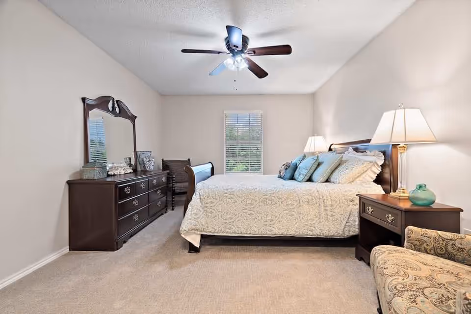 A well-lit bedroom with a large bed covered in a white patterned bedspread and multiple pillows. There is a dark wooden dresser with a mirror on the left side, a window with blinds in the center background, and two bedside tables with lamps on either side of the bed. A patterned armchair is partially visible in the foreground on the right side. The room has beige walls and carpet, and a ceiling fan with lights is mounted on the ceiling.