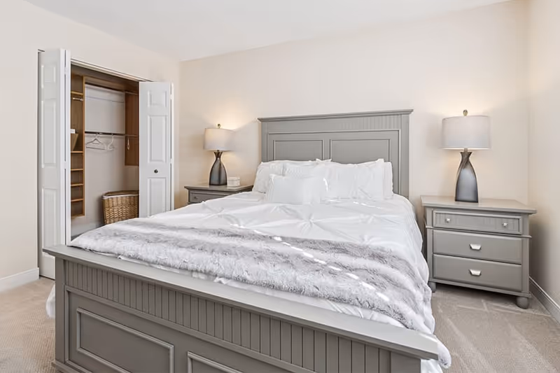 A neatly made bed with white bedding and a gray headboard in a bedroom. There are two matching gray nightstands on either side of the bed, each with a lamp. An open closet with white bi-fold doors is visible on the left side of the room, showing some hangers and a wicker basket inside. The walls are painted a light beige color.