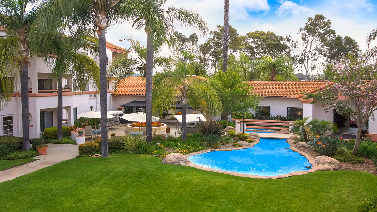 Courtyard with a small decorative pool, palm trees, patio umbrellas and seating, and surrounding two-story buildings with red tile roofs.