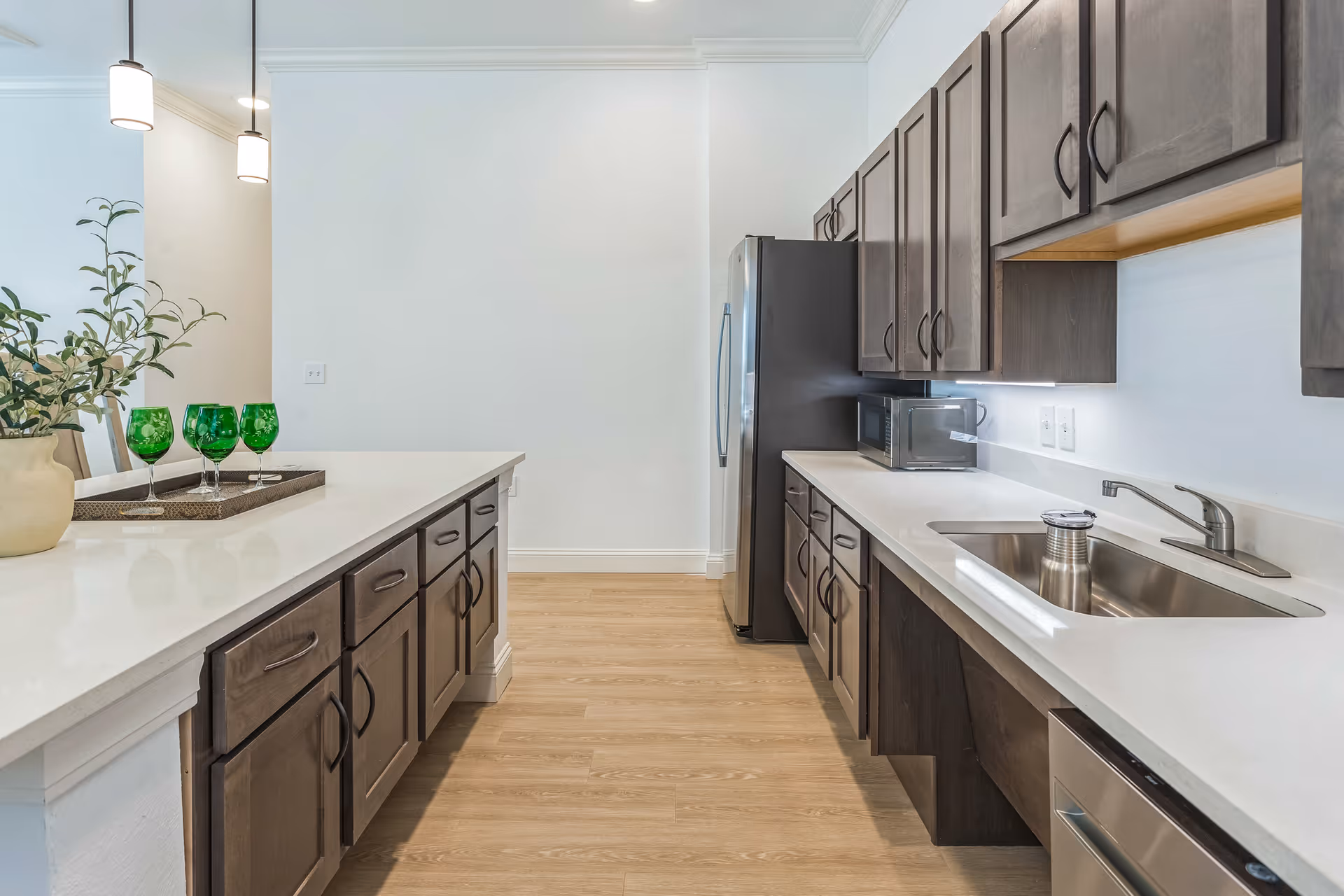 Modern kitchen with wooden cabinets, white countertops, stainless steel sink, microwave, refrigerator, and a kitchen island with a decorative plant and four green glasses on a tray.