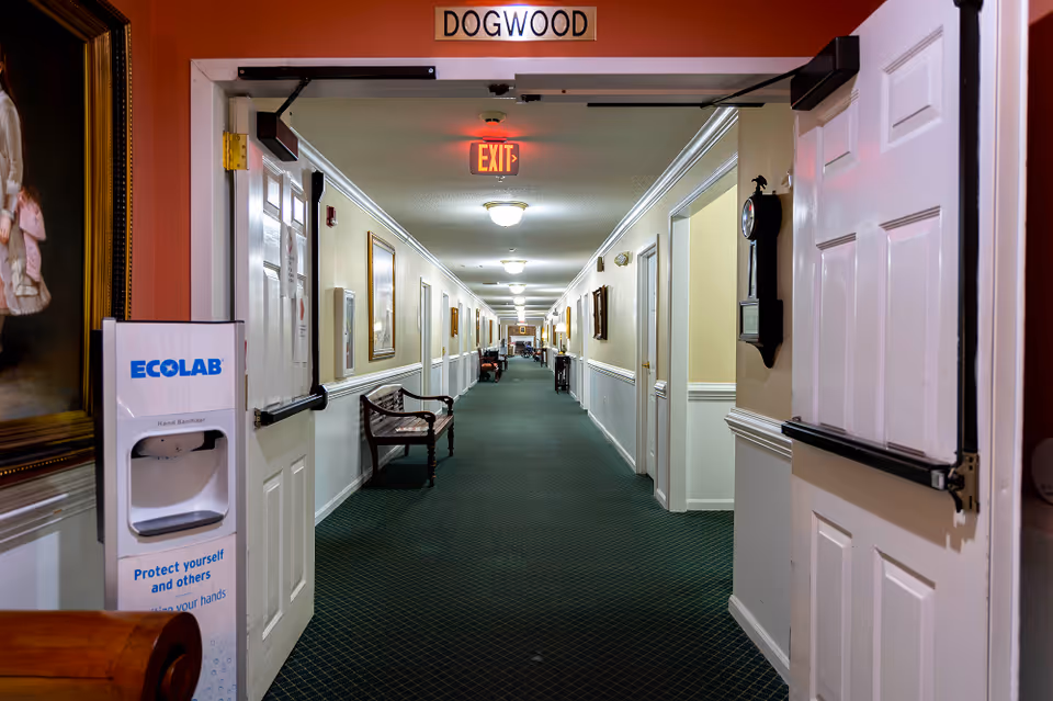 View down a long, carpeted hallway in a senior living facility named Dogwood, with white walls, several closed doors, framed pictures, benches along the walls, and an exit sign illuminated at the ceiling. A hand sanitizer dispenser is visible on the left side near the entrance.
